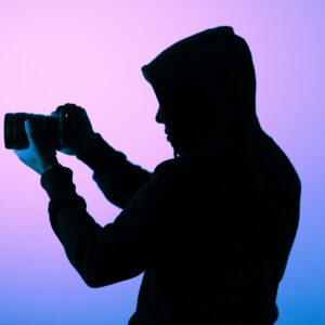 A silhouette of a hooded photographer capturing a shot with a camera in a softly lit studio.